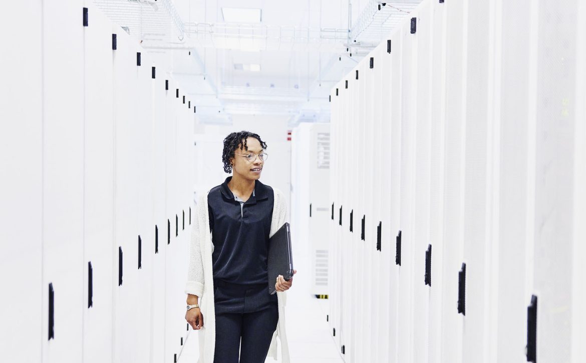 Wide shot of female computer engineer holding laptop while walking between server racks in data center