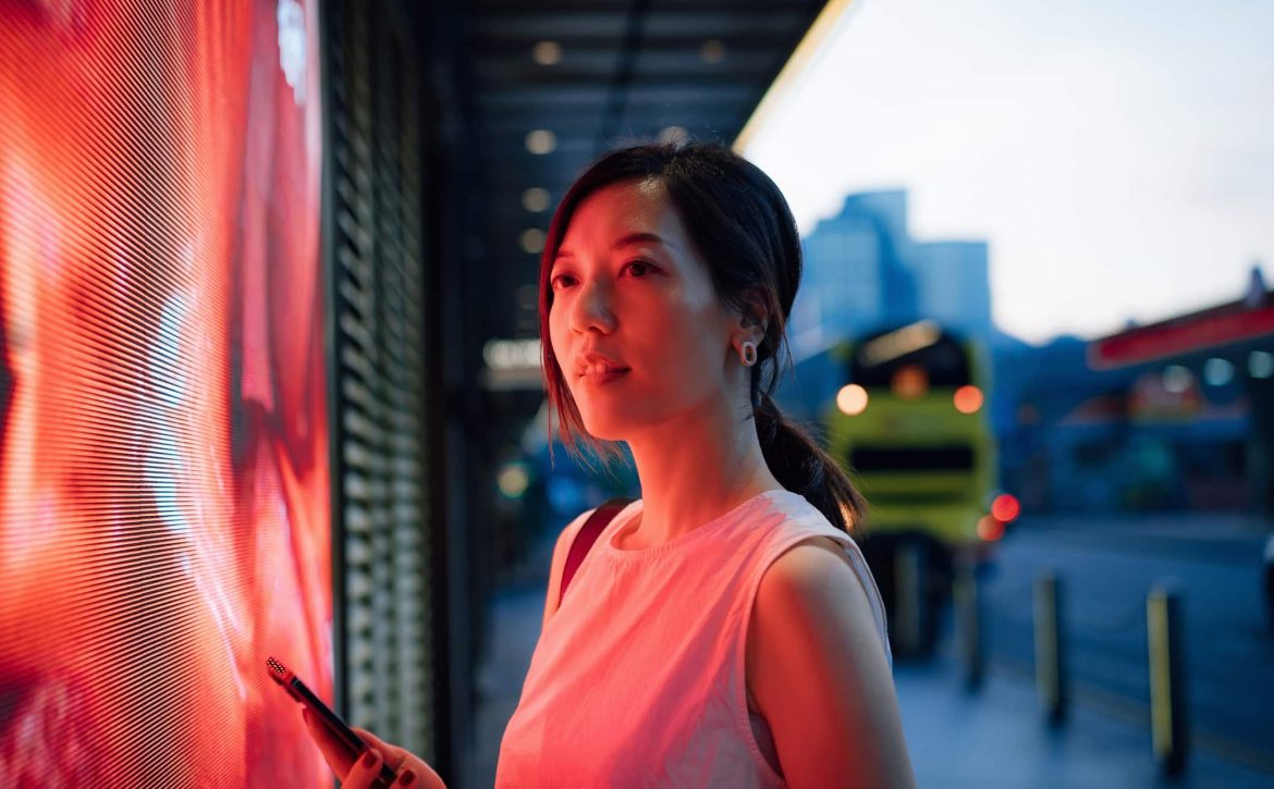 Confident young Asian businesswoman standing against illuminated LED digital display in the city, lit by red neon coloured lights, using smartphone in downtown city street in the evening. Lifestyle and technology